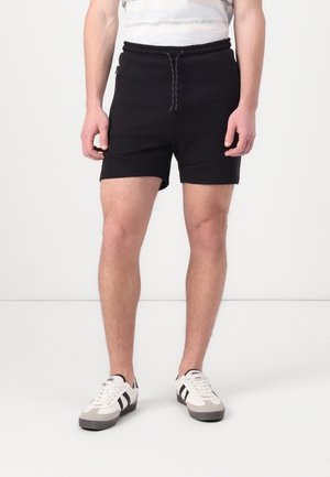 Homme portant un short noir à cordon, des baskets blanches avec des rayures noires, debout sur un sol gris clair contre un fond blanc uni.