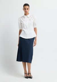 Witte blouse met pofmouwen en decoratieve versieringen; gecombineerd met een knielange donkerblauwe rok en zwarte puntige hakken.