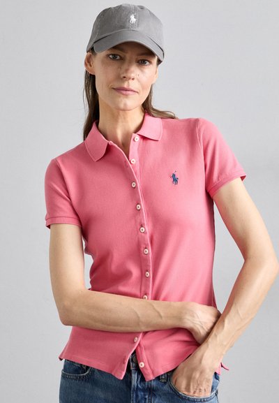 Rosa Polo-Shirt mit kurzen Ärmeln, mit einer geknöpften Vorderseite und einem kleinen gestickten Logo. Getragen mit einer grauen Kappe.