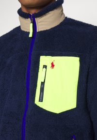 Marinblå fleecejacka med hög krage, ljusgul dragkedjeficka och kontrasterande blå dragkedjedetalj. Mjuk textur med sportig design.