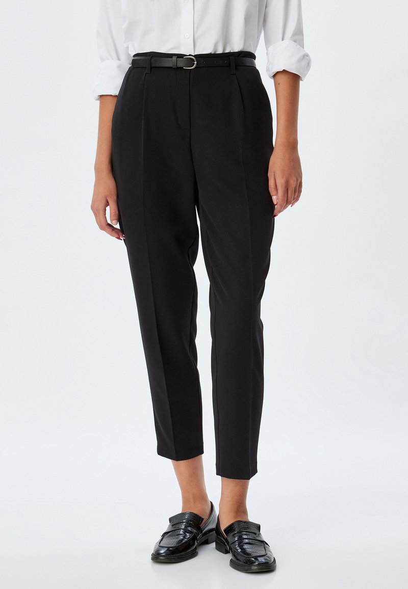 Pantaloni neri su misura a vita alta, con pieghe frontali e una sottile cintura nera. Indossati con mocassini neri e una camicia bianca.