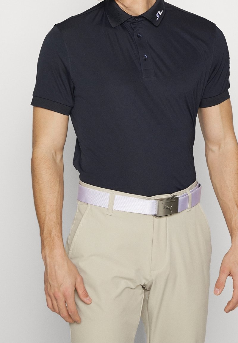 Granatowa koszulka polo z kołnierzykiem i trzema guzikami, w zestawieniu z jasnobeżowymi spodniami. Posiada jasnofioletowy pasek z metalową sprzączką.