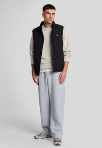 Chaleco negro con cremallera sobre un sudadera gris claro, combinado con pantalones de chándal gris holgados y zapatillas gris y verde, usado por un hombre de pie.