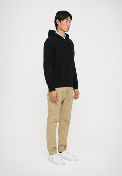 Sweatshirt preto com capuz e bolsos frontais, combinado com calças cargo bege e ténis brancos, em pé sobre um fundo neutro.