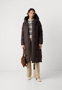 Femme portant un long manteau doudoune marron foncé sur un pull blanc à motifs, un pantalon beige, des bottes claires, et tenant un sac à main marron.