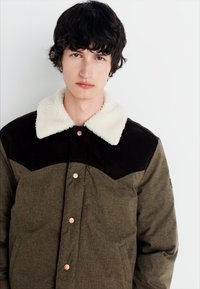 Veste vert olive avec un empiècement en suède marron et un col en fausse fourrure crème, comportant des boutons-pression et un fini en tissu texturé.