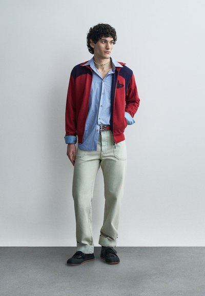 Νεαρός άνδρας που φοράει κόκκινο και μπλε μπουφάν πάνω από μπλε πουκάμισο με κουμπιά, ανοιχτό μπεζ παντελόνι, μαύρα παπούτσια, στέκεται μπροστά σε απλή γκρι τοίχο.