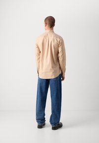Chemise boutonnée beige clair à manches longues, avec un ourlet arrondi et un pli au dos ; associée à un jean bleu ample et des chaussures noires.