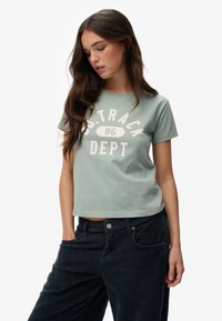 Lys grøn kortærmet T-shirt med "SD. TRACK 86 DEPT" trykt i hvid. Lavet af bomuldsstof med en afslappet pasform. Sammen med mørke fløjlsbukser.