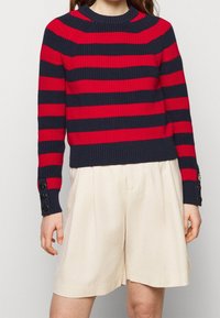 Czerwono-granatowy sweter w paski z okrągłym dekoltem i mankietami na guziki. Połączony z beżowymi szortami. Tkanina o fakturze.