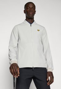Light gray zip-up jacket with a high collar, elastic cuffs, and a small yellow logo. Smooth texture, minimalist design, paired with dark pants.