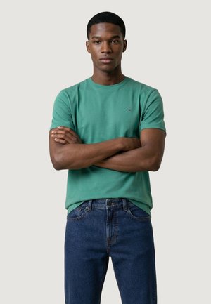Giovane uomo con le braccia incrociate che indossa una maglietta verde a maniche corte e jeans blu, in piedi contro uno sfondo chiaro e semplice.