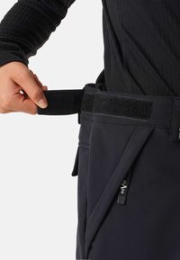 Czarne spodnie softshellowe z regulowanym pasem na rzepy, z kieszeniami i gładką teksturą. Ręka chwytająca pas w celu regulacji.