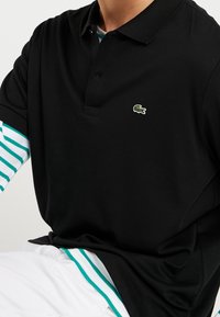 Polo noir avec un logo crocodile vert sur la poitrine, manches courtes avec un motif rayé blanc et turquoise en dessous.