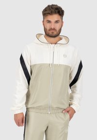 Leichte, zweifarbige Jacke: obere Hälfte in Creme, untere Hälfte in blassem Oliv, mit schwarzen Seiteneinsätzen, Reißverschluss, verstellbarer Kapuze und elastischen Bündchen.