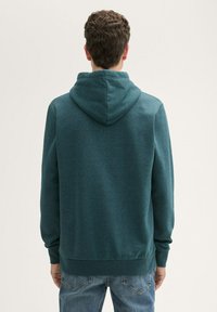 Hoodie turchese con finitura testurizzata, dotato di cappuccio con cordino, polsini e orlo a coste. Indossato con jeans azzurri, visto da dietro.