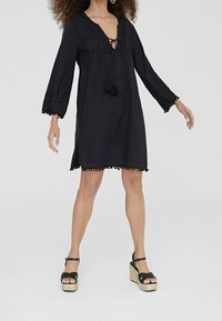 Svart tunikaklänning med långa ärmar, broderade detaljer, knytkrage, fransar och pom-pom-accents vid ärmslut och nederkant. Bärs tillsammans med platåsandaler.