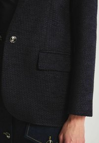 Blazer texturé bleu marine à coupe droite, avec une poche poitrine simple et des boutons argentés brillants. Il présente un motif tissé subtil et des manches structurées.