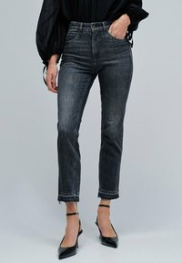 Jeans de mezclilla negra con una cintura alta, diseño de pierna recta y dobladillo deshilachado. Llevados con tacones de aguja negros y puntiagudos, y una blusa negra holgada.