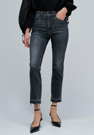 Jeans de mezclilla negra con una cintura alta, diseño de pierna recta y dobladillo deshilachado. Llevados con tacones de aguja negros y puntiagudos, y una blusa negra holgada.