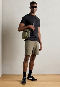 Sort t-shirt, olivengrønne shorts, sorte sportssko med hvide striber og en lille olivengrøn skuldertaske. Model iført mørke solbriller.