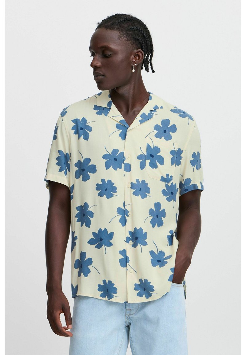 Cămășă cu mânecă scurtă, dintr-un material galben deschis, cu modele florale de culoare albastru închis. Design fără guler, cu un buzunar pe piept și o croială lejeră.