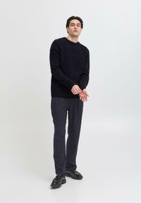 Pulover tricotat călduros, de culoare albastru marin, cu guler rotund, manșete și tiv cu dungi, asortat cu pantaloni închiși la culoare și pantofi negri. Design și textură simple.