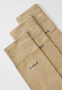 Bambuske do koljena od bež rebrastog pletenog materijala, s čvrstim manžetama i izvezenim "BOSS" logotipom u tamnoplavoj boji sa strane.