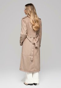 Beżowy płaszcz typu trencz z paskiem w talii, pagonami na ramionach, długimi rękawami i sięgający do kolan, zestawiony z białymi spodniami o szerokich nogawkach.