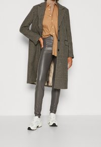 Lång svartvitt houndstooth-kappa över en beige skjorta med knappar, grå läderleggings och vita sneakers med färgglada accenter.