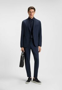Completo blu navy su misura con una camicia abbottonata coordinata, caratterizzato da una texture liscia; accessorato con una borsa in pelle nera e sneakers scure.