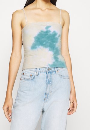 Frau trägt ein hellblaues Tie-Dye-Spaghetti-Top in Kombination mit hoch taillierten hellblauen Jeans und minimalistischem Goldschmuck in Form von Ringen.