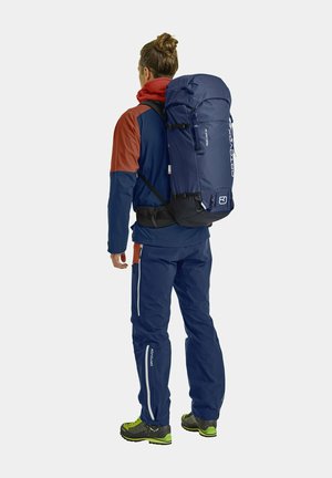 Rückansicht einer Person, die eine blau-orange Jacke, eine marineblaue Hose und einen großen blauen Rucksack mit weißer Markenbeschriftung und obenem Verschluss trägt.