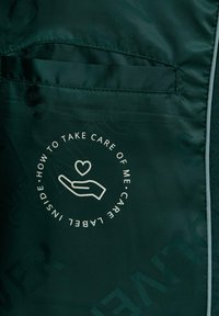 Tissu vert foncé avec une étiquette d'entretien imprimée montrant une main tenant un cœur et le texte "How to take care of me care label inside."