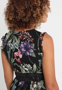 Robe fleurie avec un fond noir orné de fleurs colorées, accents à volants sur les épaules, et une ceinture noire en crochet.
