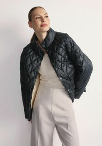 Pikowana czarna kurtka puchowa z błyszczącego materiału, z wysokim kołnierzem i zatrzaskami, częściowo rozpięta na jasnej bluzce, w połączeniu z jasnymi spodniami.