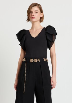 Femme portant un haut noir à col en V avec des manches bouffantes superposées, un pantalon noir taille haute et une ceinture chaîne dorée ornée de médaillons circulaires.