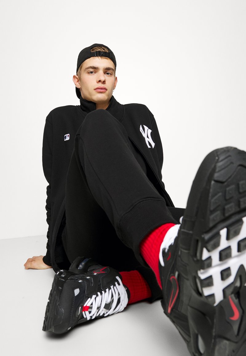 Black athletic jacket with white logo, matching black pants, red and white socks, and black sneakers with white laces and accents.