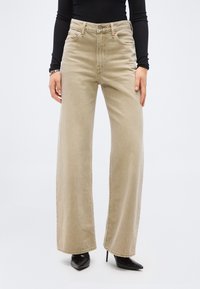 Μπεζ τζιν με ψηλή μέση και φαρδιά μπατζάκια, φορεμένα με μαύρα ψηλοτάκουνα παπούτσια με μυτερή μύτη και μαύρο τοπ με μακριά μανίκια, τα χέρια χαλαρά στα πλάγια.