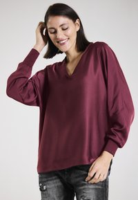 Burgundy, silky blouse with a V-neck, wide sleeves, and relaxed fit. Worn with distressed black jeans; smooth, reflective texture.