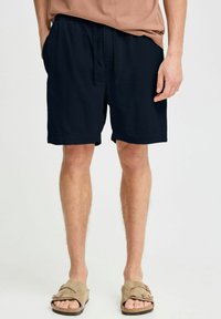 Navy blue, textured shorts with a relaxed fit and side pockets. Worn with brown sandals, paired with a light brown shirt.