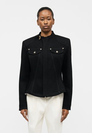 Frau trägt eine eng anliegende schwarze Jacke mit knöpfbaren Brusttaschen und weißen Nähten, kombiniert mit lockeren weißen Hosen, steht vor einem schlichten Hintergrund.