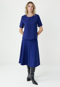 Kobieta ubrana w granatowy krótki rękaw i dopasowaną midi spódnicę, w czarnych kozakach sięgających do kolan, stojąca na jednolitym tle.