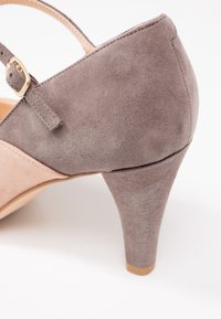Gros plan sur une chaussure à talon haut en daim taupe, dotée d'un talon arrondi et d'une bride à la cheville avec une petite boucle dorée.