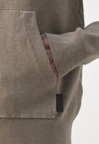 Main dans la poche avant d'un pull en tricot taupe avec poignets côtelés et petite étiquette en tissu noir sur la couture latérale.