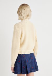 Pull en tricot crème avec texture côtelée et col montant, associé à une jupe en denim ornée de broderies de papillons rouges.