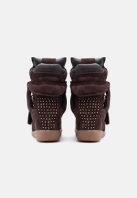 Ζευγάρι σκούρων καφέ σουέντ αθλητικών παπουτσιών με χρυσές λεπτομέρειες από τρουκς στη φτέρνα και λαστιχένιες σόλες χρώματος καλαμποκιού, από πίσω.