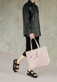 Camicia nera in pelle con bottoni, gonna nera plissettata, borsa a tracolla rosa in vernice con dettaglio a fiocco e sandali neri con plateau e cinturini regolabili.