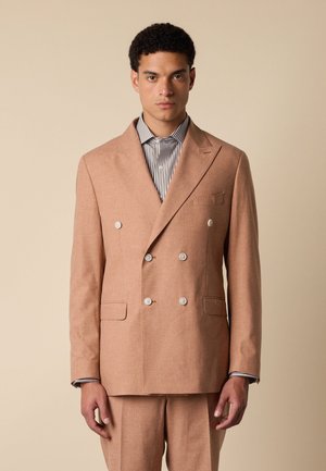 Homme portant une veste de costume croisée beige et une chemise rayée, debout devant un fond beige uni, face à nous.