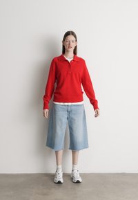 Červený pletený polo sveter s golierom, dvoma gombíkmi a rebrovanými manžetami, kombinovaný s denimovými šortkami po kolená vo svetlom umývaní a bielymi teniskami.